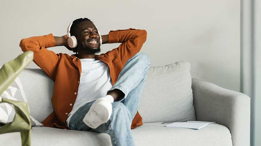 Mann sitzt auf Sofa hört Audio und schaut strahlend aus dem Fenster.