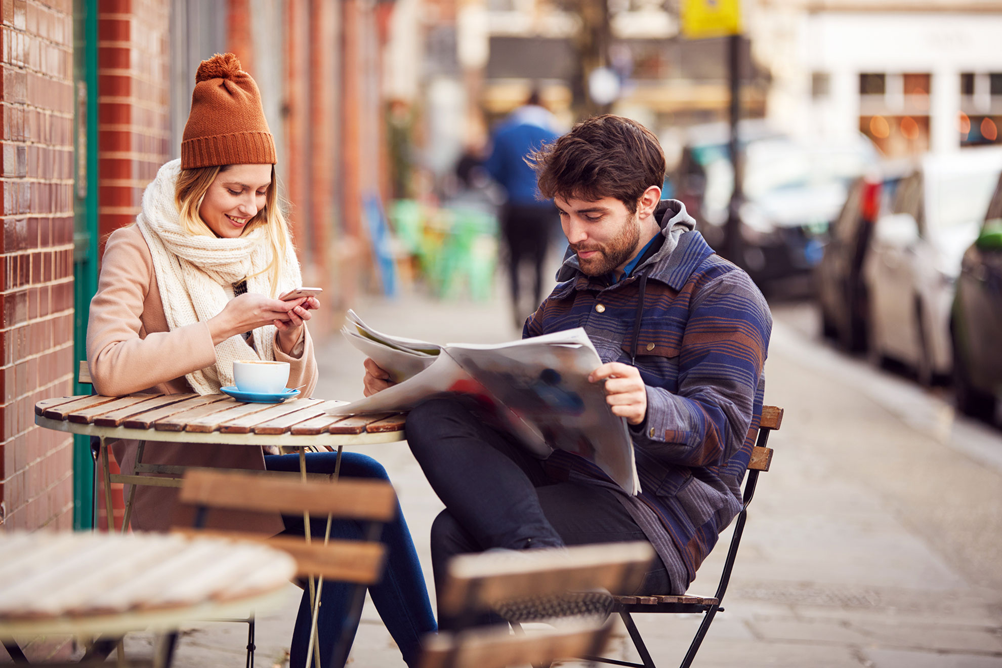 Zwei junge Leute sitzen an in Klamotten eingemurmelt vor einem Café lesen Zeitung und schauen aufs Mobile.