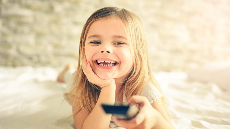 Kind das glücklich auf dem Bett liegt mit einer Fernbedienung in der Hand und TV schaut.