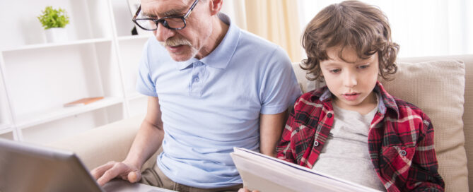 Opa und Enkel sitzen auf dem Sofa. Der Opa mit dem Laptop auf dem Schoss, während der Enkel die Zeitung liest.