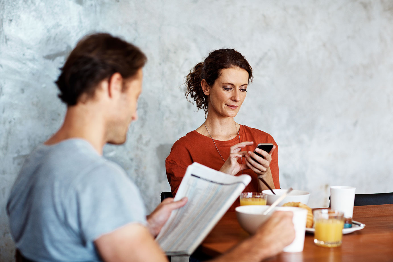 Frau und Mann sitzen am Tisch und lesen, er Zeitung, sie am Handy.